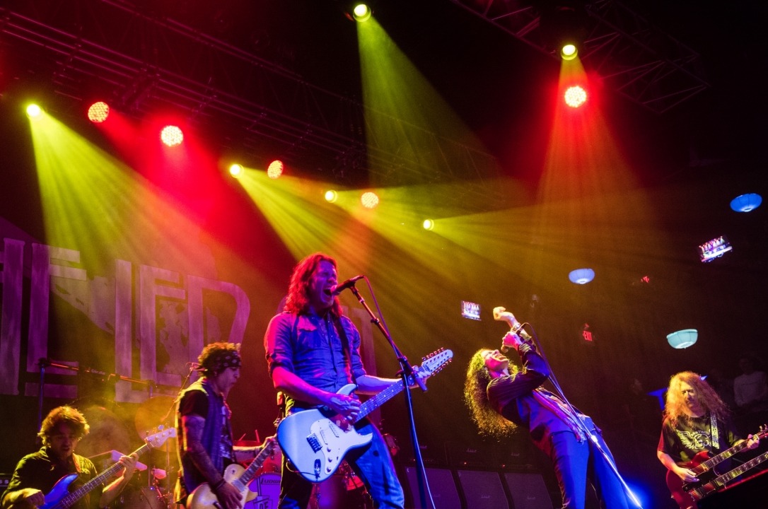 A man with long hair singing while playing guitar stands with two guitar players to his left, a long haired man leaning back with his arm in the air singing into a microphone and a man playing guitar to his left underneath red and green stage lights.