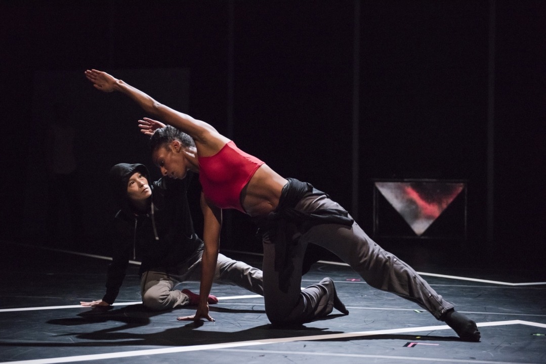 Woman in red top and grey pants on one hand and one knee reaching over her head with a person in black behind her mimicking her all in front of a black background.