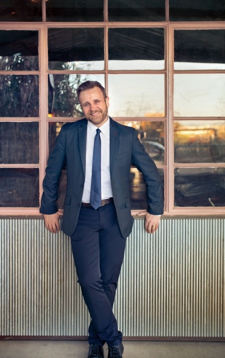 A man in a blue suit and tie and a white button down standing in front of a window and smiling at the camera.