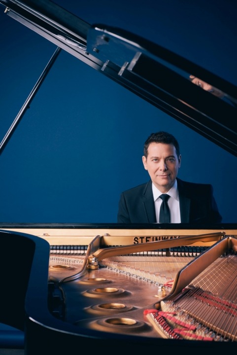 Michael Feinstein in a black suit and black tie and a white shirt sitting behind an open grand piano looking at the camera in front of a blue background.
