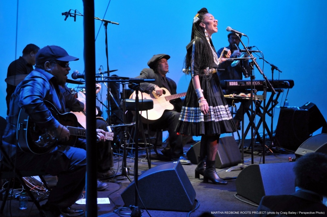 Woman wearing a black dress singing into a microphone while playing a percussion instrument with five other people in black playing instruments around her in front of a blue background.