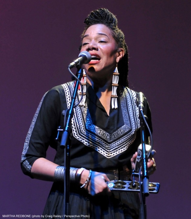 Woman wearing a black top singing into a microphone while playing a percussion instrument in front of a purple background.