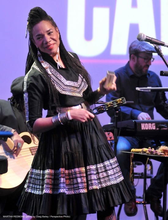 Woman wearing a black dress and smiling while playing a percussion instrument and a man in black playing the keyboard behind her all in front of a purple background.