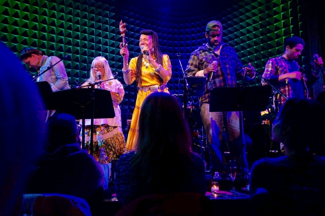 A woman in a yellow dress singing and holding an instrument with four background singers behind her all in front of a green background.