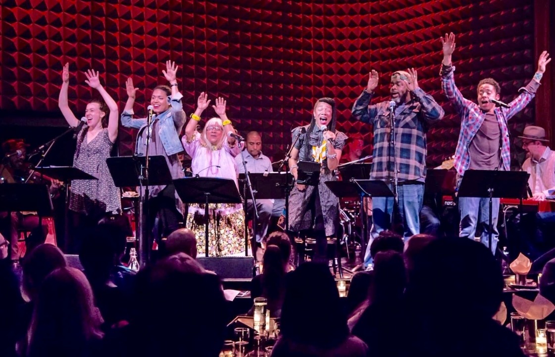 6 people singing and 3 people seen playing instruments behind them. Everyone is on a sage in front of a red background.