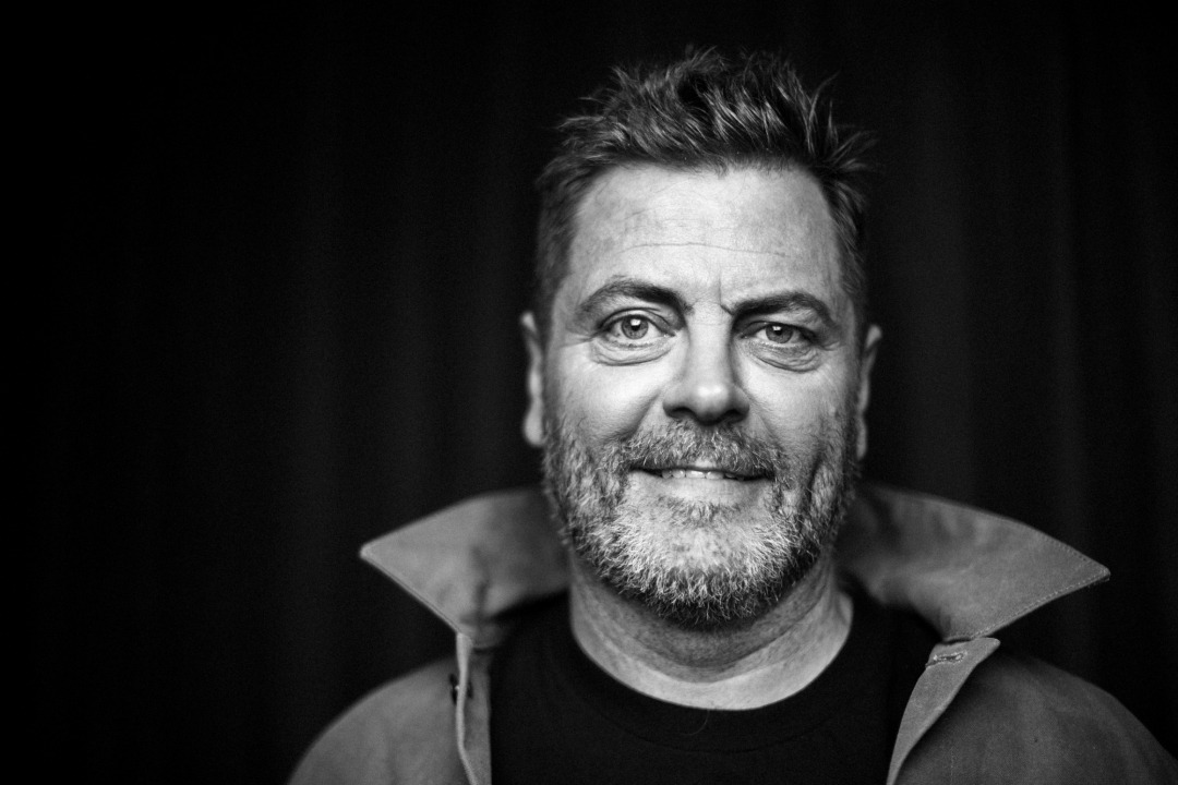 Black and white photo of Nick Offerman in a black shirt and a coat with a popped collar in front of a black background.