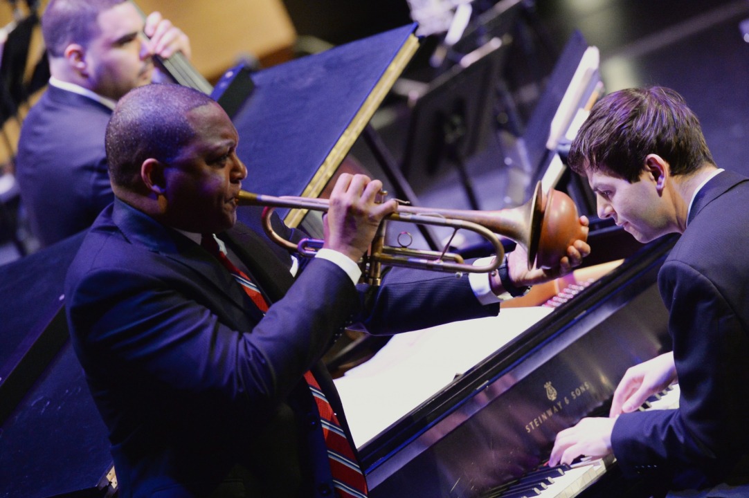 Male african american, wearing a black suit with a red, white, and blue striped tie, is playing a trumpet with a plunger. White man playing the piano to the trumpet player's left. And a male cello player is also visible behind the trumpet player.