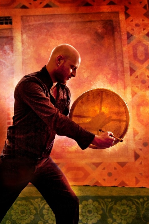 A man in a black shirt and ants playing a drum in front of an orange and green background.