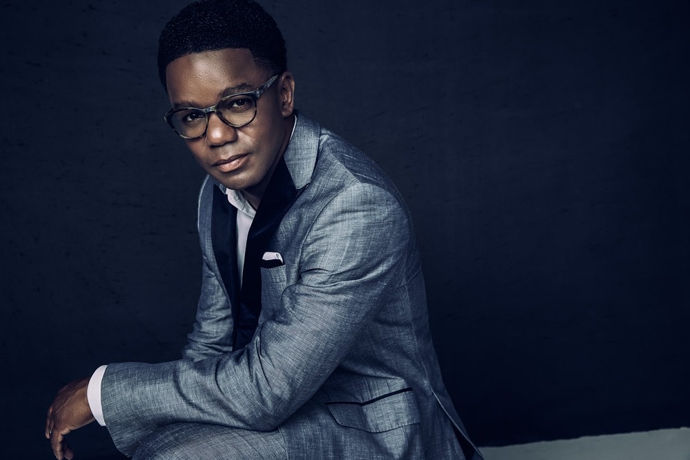 An African American man wearing glasses sits in front of a dark blue background wearing a gray suit.