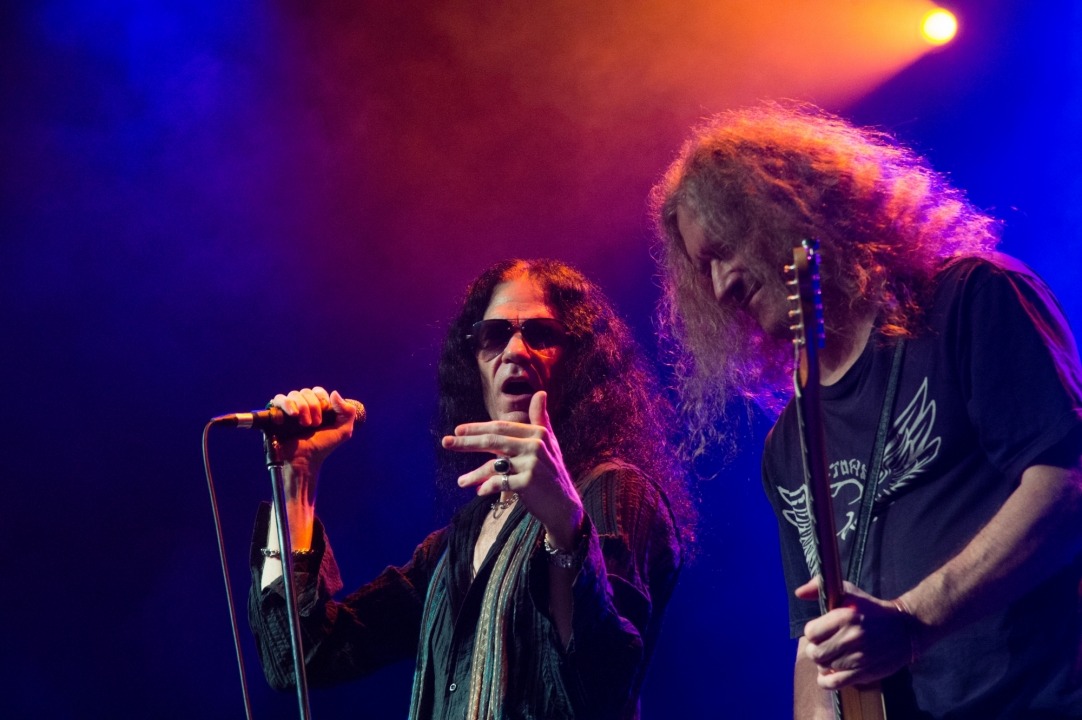 Two men stand together, the one on the left has long dark hair and sunglasses on, holding a microphone and looking towards the man on the right. The man on the right has long blonde hair, is wearing a black t-shirt, is playing guitar and is looking down.