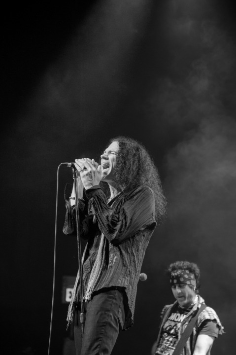 A black and white photo of a man with long dark hair singing into the microphone while a man behind him plays guitar.