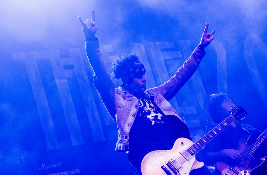 A blue tinted photo of a man with an electric guitar standing with his arms in the air while another guitar player plays behind him.