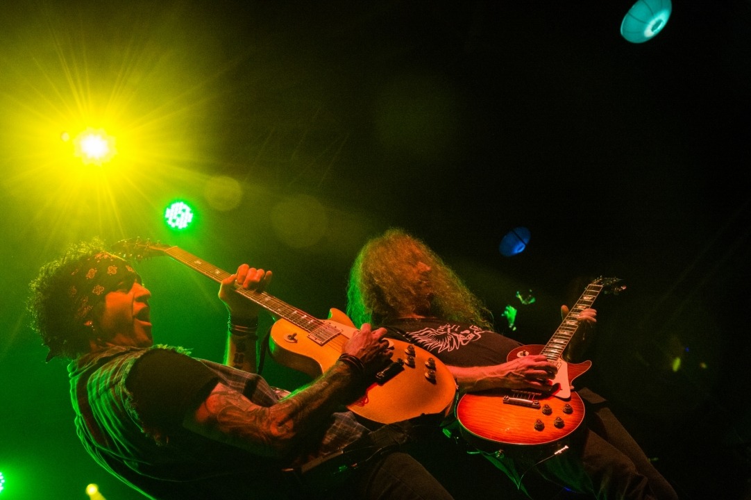 Two men wearing all black stand together playing electric guitars with yellow and green lights shining behind them.
