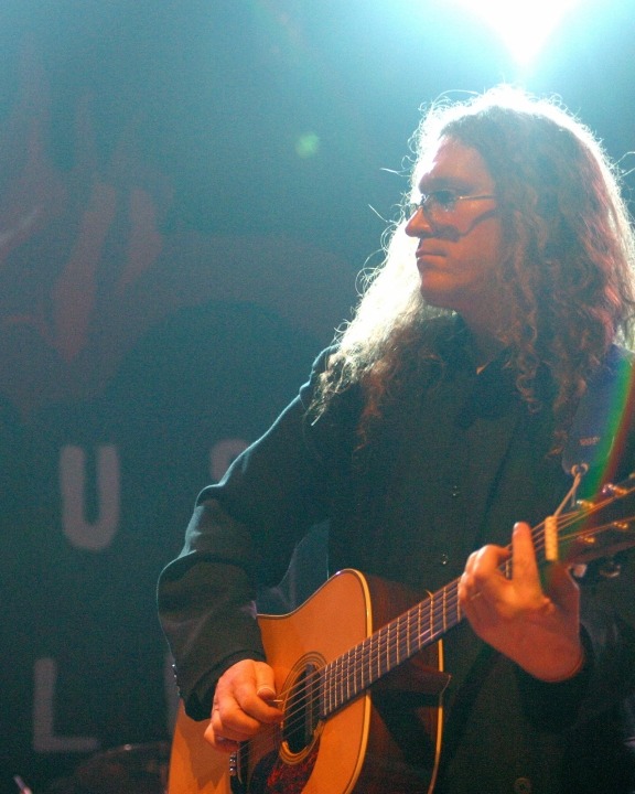 The side view of a man with long hair and glasses stands in a black button down shirt playing an acoustic guitar.