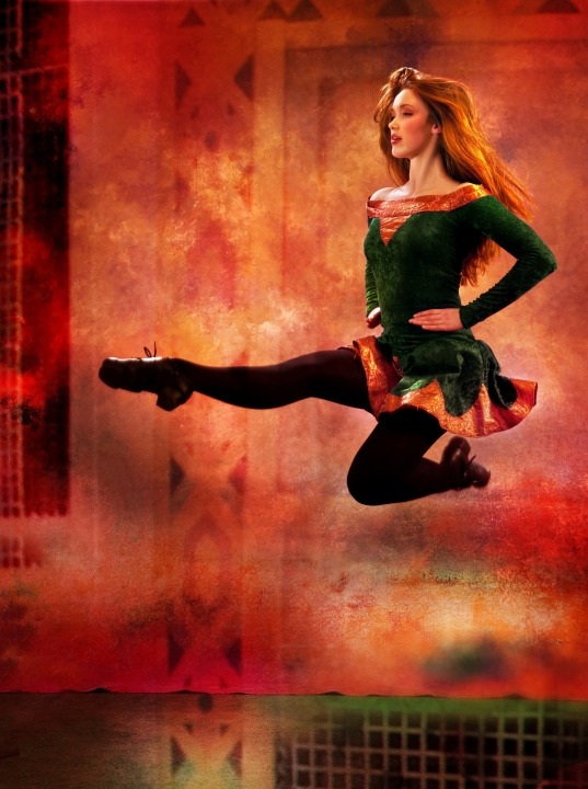 A woman in a green dress and black tights Irish dancing in front of an orange background.