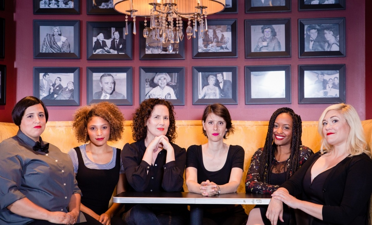 Six women in black clothing sitting at a table on a yellow couch with a red wall full of black and white pictures behind them.