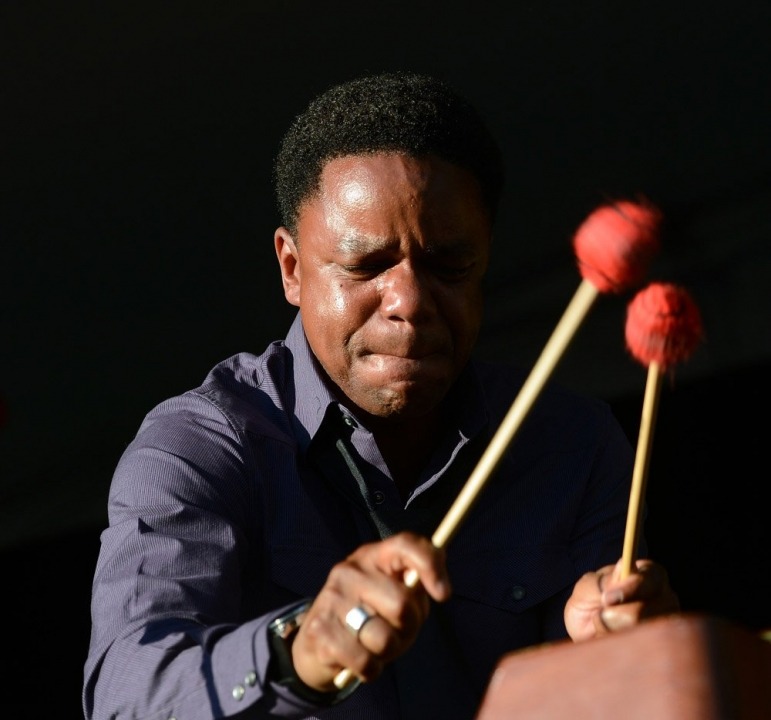 An African American man holds two drumsticks with red ends in front of a black background with his eyes closed.