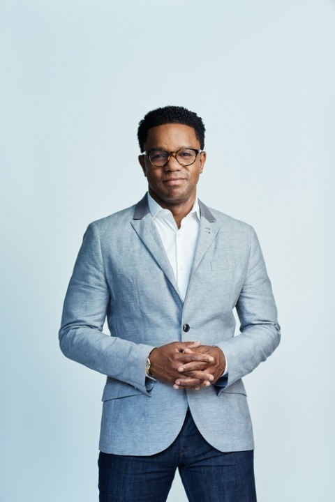 African American man wearing glasses in front of a bright blue background stands with his hands folded wearing a light gray suit jacket.