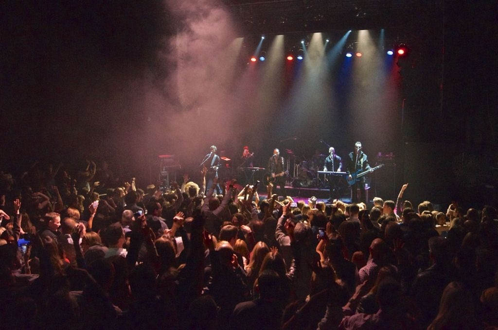 Six band members wearing black on stage playing instruments in front of a crowd of people all in front of a black background.