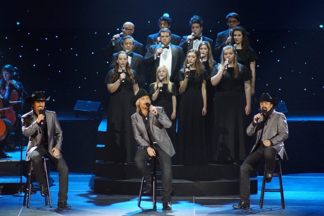 Three men sitting on stools singing into handheld microphones wearing black cowboy hats, black button down shirts, grey suit jackets, and black jeans stand in front of a choir of 12 college aged men and women standing on risers wearing long black dresses and tuxes singing into microphones. They are all in front of a black background with twinkling lights.