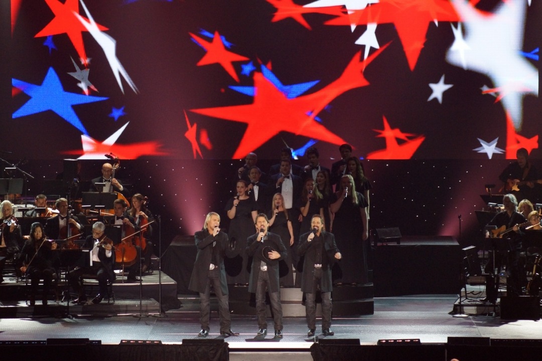 Three men standing wearing black button down shirts, thigh length black suit jackets, and black jeans sing into handheld microphones while standing in front of a choir of 12 college aged men and women standing on risers wearing long black dresses and tuxes singing into microphones. There is an orchestra of cello players to the left of them and violin players to the right. They are all in front of a background with red white and blue stars projected onto it.