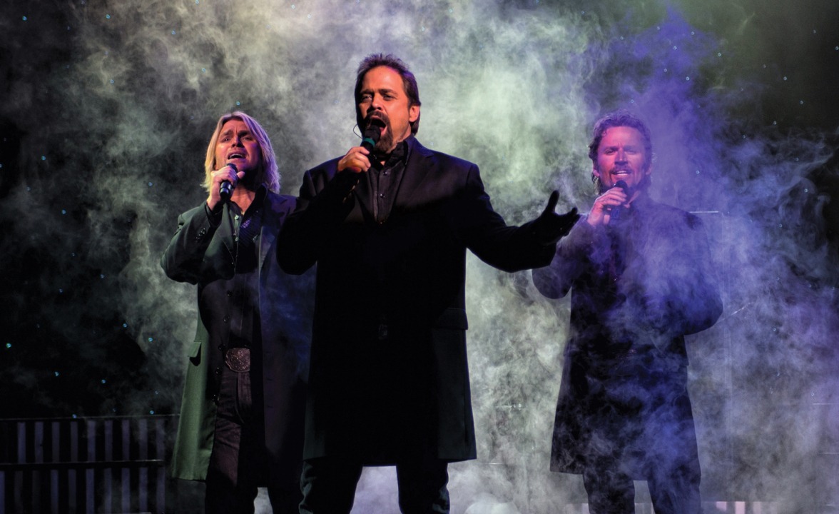 Three men stand in front of a black smoky background singing into microphones. They are wearing black button down shirts and thigh length black suit jackets.