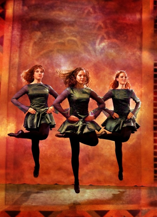 Three women Irish dancing in green dresses and black tights in front of an orange background.