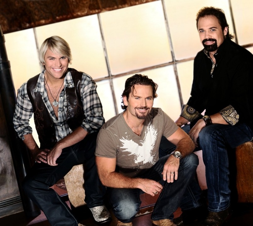 Three men sit posed. On the left, a blonde wearing jeans, a black and white plaid shirt and a brown leather vest, the middle one a brunette squatting below the two wearing jeans and a brown v-neck tshirt with a white splatter on the front and the one on the right is a brunette sitting while wearing a black button down and jeans.
