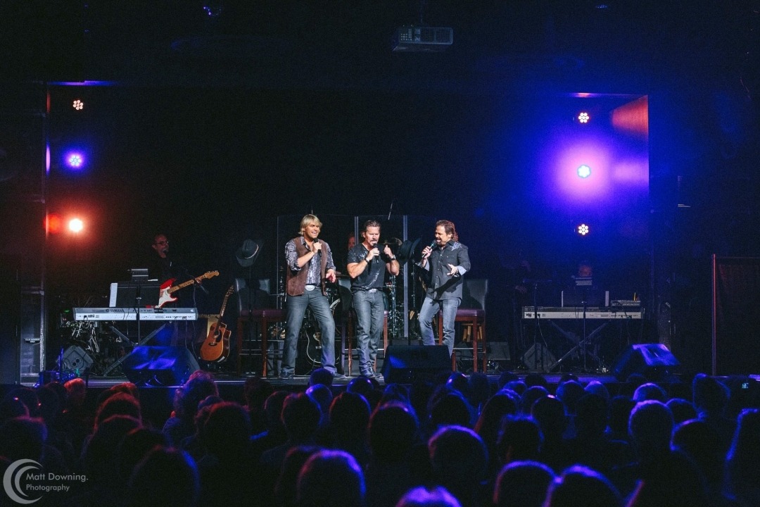 Three men stand on a stage in front of an audience wearing jeans and black button down shirts singing into handheld microphones with two keyboards on either side of them , a guitarist behind them and a black background with multicolored lights lining the left and right side of the stage.