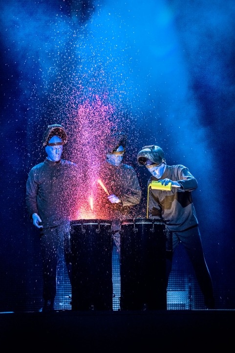 Three bald blue man standing in front of two drums pouring paint onto them in front of a blue and black background