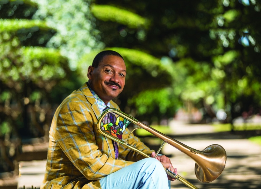 Delfeayo Marsalis stitting outside wearing a yellow suit jacket and light blue pants and holding his trombone