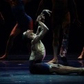 Image of a male performer with no shirt and black shorts seated on the stage with hands raised looking toward the sky while the legs of other performers are visible in the background.