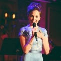 A woman in a light blue dress with a microphone smiling into the camera