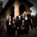 Image from a point of view looking up at the eight ukulele players who are standing outside on steps and holding their ukuleles while looking in various directions with pillars in the background