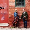 Image of the two performers from Celtic Celebration holding hands and their fiddles while standing in front of a brick wall.