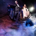 Phantom in white mask and black suite holding a lantern and leading Christine, who is wearing a white dress, down stone steps with a dark background and fog beneath them.