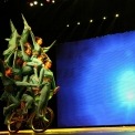 Eight acrobats wearing green costumes riding a single bicycle with a blue light background.