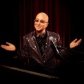 Paul Shaffer posed behind his keyboard and microphone wearing a sequin suit and shrugging to the camera