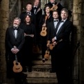 Image of the eight ukulele players holding their ukuleles and posed on steps smiling into the camera with stone walls behind them