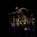 Image of a shirtless male performer being held aloft by multiple male and female performers.