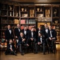 Ten men in blue velvet suits and bow ties sitting with each other and laughing with a grand bookcase in the background