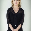 Image of Gabriela Monteri in a black shirt standing in front of a white background and staring at the camera with hands clasped at her waist.