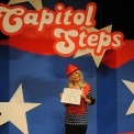 Actor dressed up as Kellyanne Conway wearing a bright red hat and holding a sign that says "Alternative Facts." She is standing in front of a red, white and blue background with Capitol Steps displayed overhead