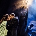 Christine, wearing a green cloak, and Raul, wearing a black coat almost kissing in front of a foggy, dark background with statues behind them.