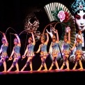 Nine women in blue costumes performing a synchronized Chinese yo-yo routine with a Chinese woman on a screen in the background