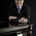 Emanuel Ax stands behind his grand piano and leans forward on it while he smiles at the camera
