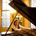 Susan Werner posed behind her grand piano looking into the camera with a tall wall and window behind her