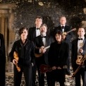 Image of the eight ukulele players holding their ukuleles standing and looking into the camera with exposed brick in the background