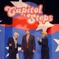 Two actors dressed up as Bernie Sanders and Hillary clinton acting upset at a third actor dressed as Donald Trump; all standing in front of red, white and blue background with Capitol Steps displayed overhead.