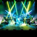Band members performing on a stage with vibrant green, yellow and blue stage lights and fog on the floor